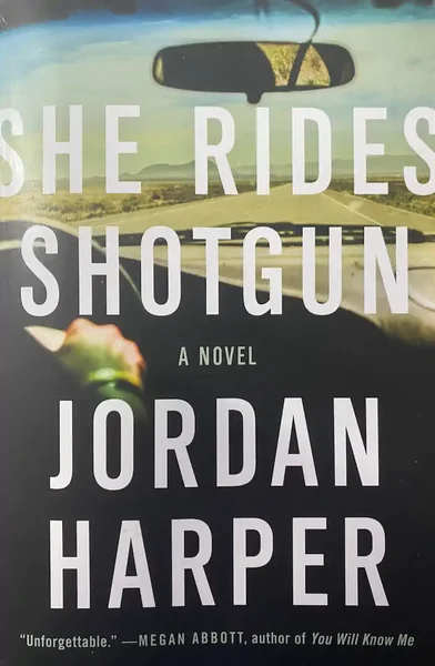 The cover of the novel "She Rides Shotgun" by Jordan Harper, showing a blurred car interior and road, with large white text displaying the title and author.
